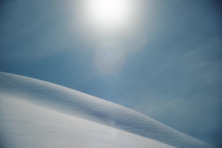 Wind blows across a pristine, sunlit snowdrift under a clear blue sky