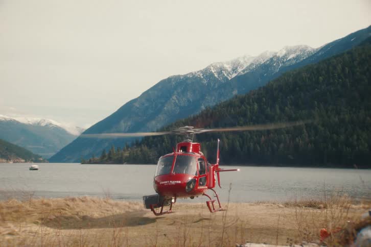 A red helicopter takes off in slow motion from the shore of a lake with majestic snow-capped mountains in the background