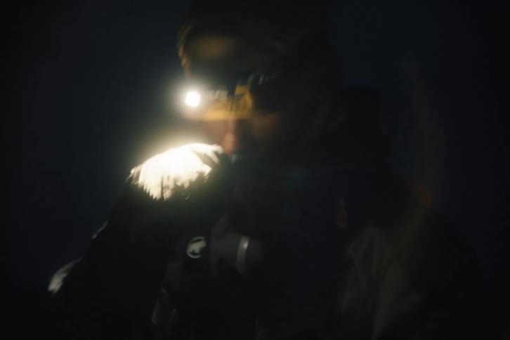 A mountaineer in a hooded jacket turns on a headlamp and adjusts equipment in the dark