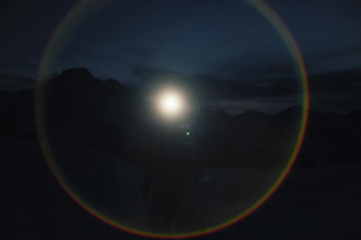 A lone splitboarder navigates a dark, snowy mountain landscape at night, their powerful headlamp creating a dramatic lens flare as they walk toward the camera