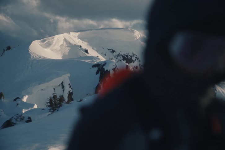 An out-of-focus hiker walks out of the frame, revealing a majestic, snow-covered mountain peak under a dramatic, cloudy sky