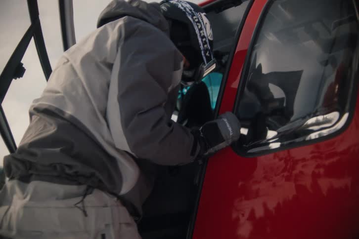 A fully-geared skier exits a red helicopter, grabs their duffel bag, and walks off into the snowy mountain backcountry