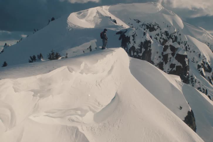 Slow zoom towards a lone skier standing on a majestic, snow-covered mountain peak, taking in the epic view