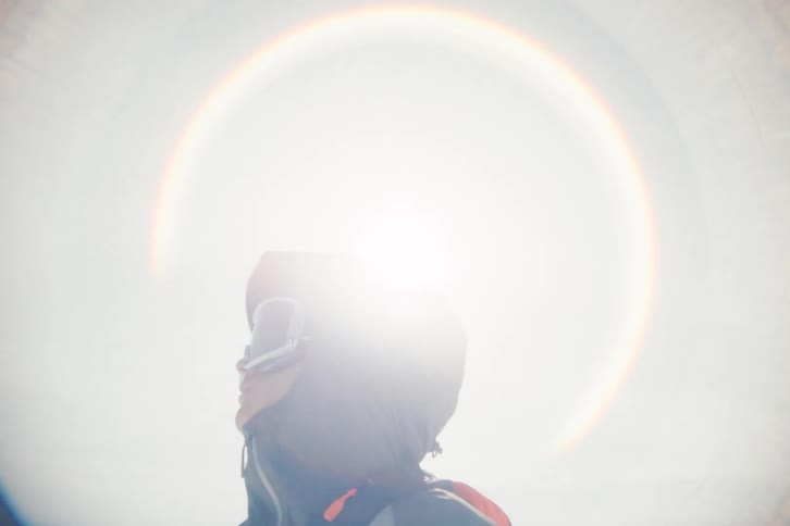A backcountry skier in a hooded jacket and goggles gazes toward the bright sun, creating a mesmerizing circular rainbow lens flare halo
