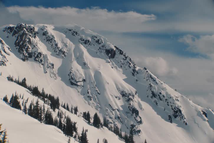 A small skier makes his way down majestic snow-covered mountain peak stands against a blue sky with slowly moving clouds