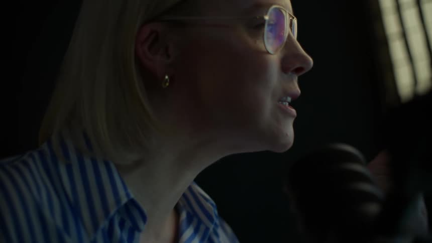 A woman with glasses and blonde hair passionately records a podcast or live stream, speaking and gesturing into a professional microphone in a dimly lit room
