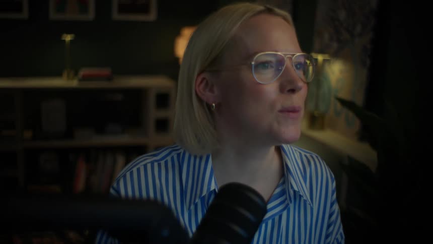 Slow zoom in on a smiling female podcaster with glasses speaking into a microphone in a cozy home studio at night