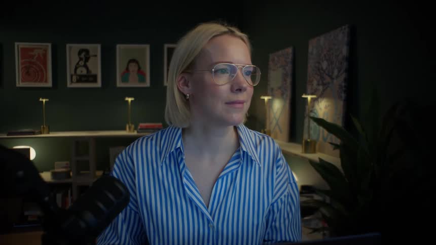 A stylish woman with blonde hair and glasses smiles while working on her laptop late at night in her cozy home studio