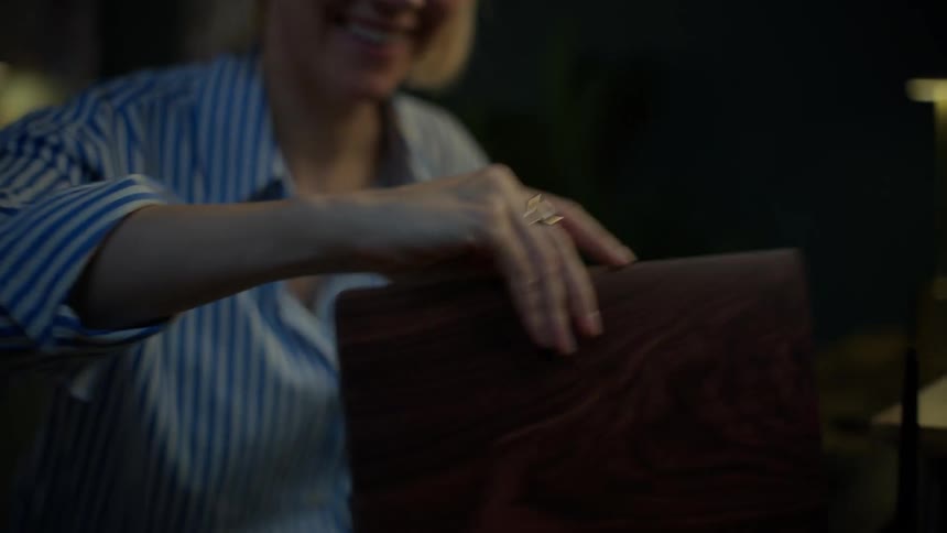 A smiling woman wearing glasses happily opens her laptop to start working late at night in her cozy home office