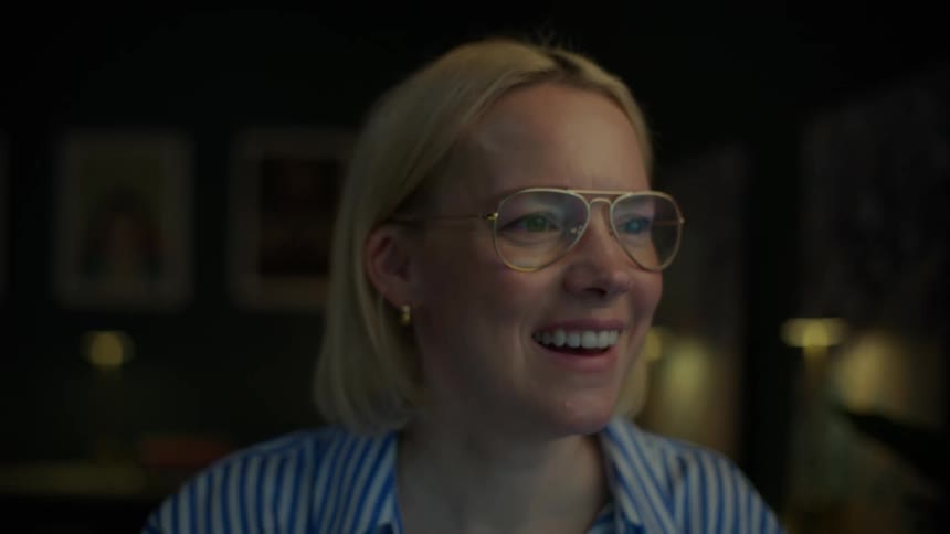 A blonde woman with glasses smiles and laughs while working on her laptop at night, with the camera slowly pulling back to reveal her home office