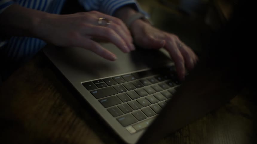 A close-up, high-angle view of a woman's hands typing on a laptop keyboard in a dimly lit room