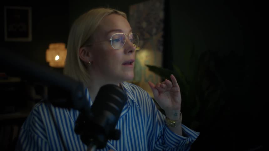 A woman in a striped shirt and glasses passionately records a podcast in her stylish, dimly-lit home office