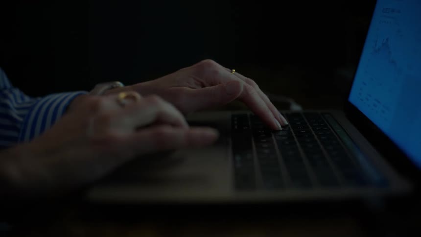Slow-motion close-up of a woman's hands typing on a laptop keyboard in a dark room illuminated by the screen's glow