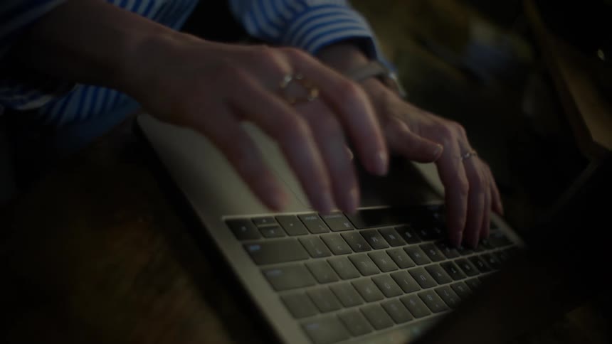 High-angle close-up shot of a woman's hands typing on a laptop in a dimly lit room