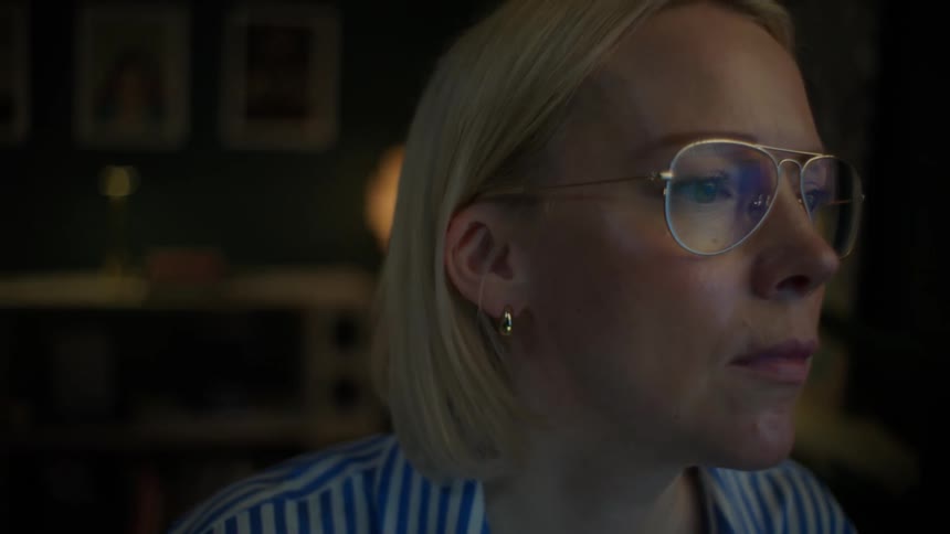 A focused woman wearing glasses working late at night in her home office is surprised and then delighted by what she sees on her computer screen