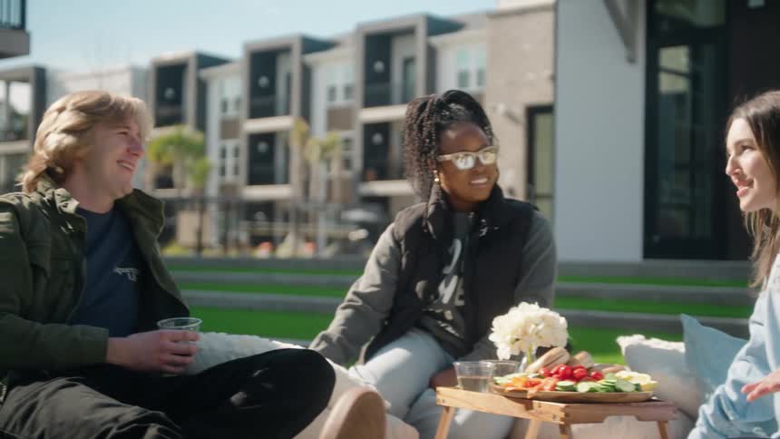 A diverse group of young friends shares laughter and conversation during a sunny outdoor picnic in a modern residential courtyard