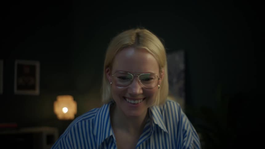 A happy blonde woman with glasses laughs while looking at her laptop screen in a cozy, dimly lit home office at night