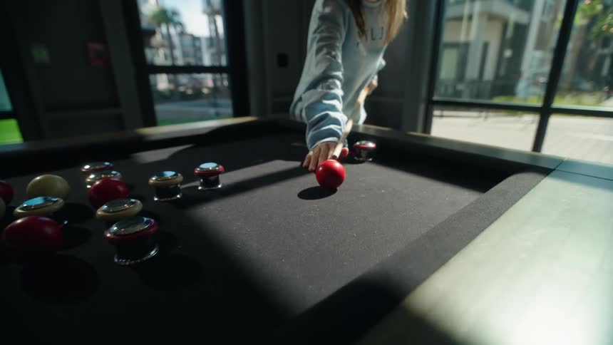 A woman carefully lines up and takes a shot with a pool cue, hitting a red puck in a slow-motion game of bumper pool