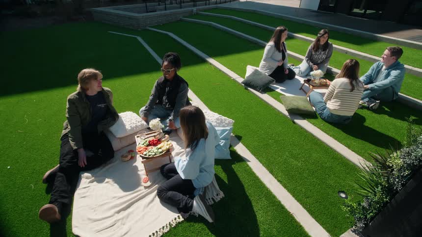 Two groups of diverse young adults socialize and enjoy a picnic on the tiered turf of a modern outdoor amphitheater on a sunny day