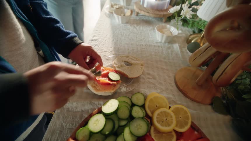 A person carefully assembles a fresh bagel with cream cheese, tomato, salmon, and cucumber from a brunch spread in slow motion