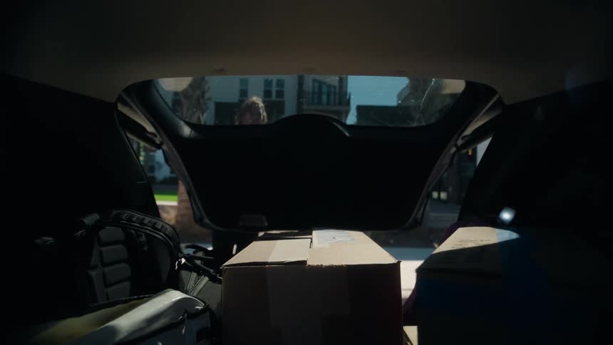 Friends greet and help unload a car full of boxes for moving day at a sunny apartment complex, seen from the trunk