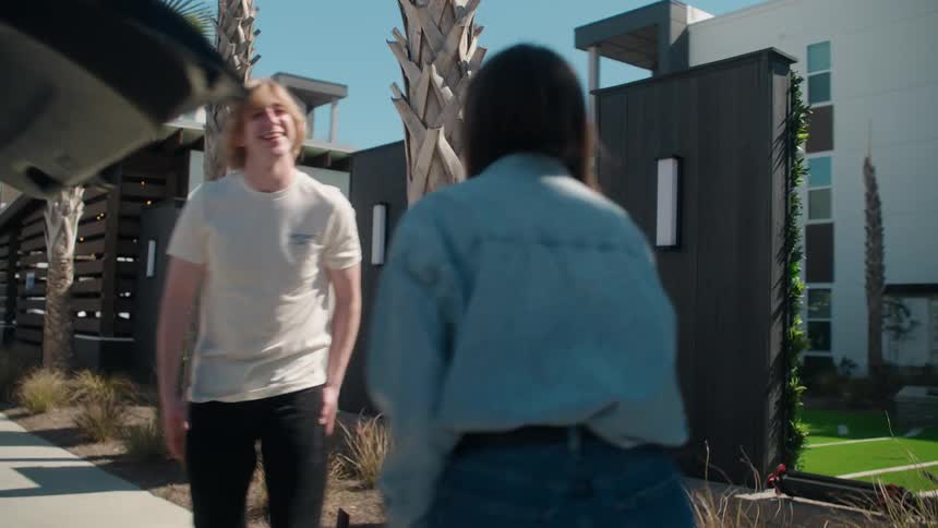 A group of young friends greets each other in an apartment complex parking lot and begins unloading luggage from the back of a car
