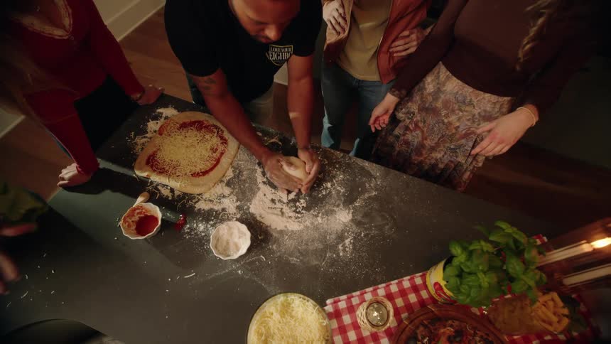 A group of friends watch as a man kneads pizza dough on a floured countertop in this slow-motion, top-down shot