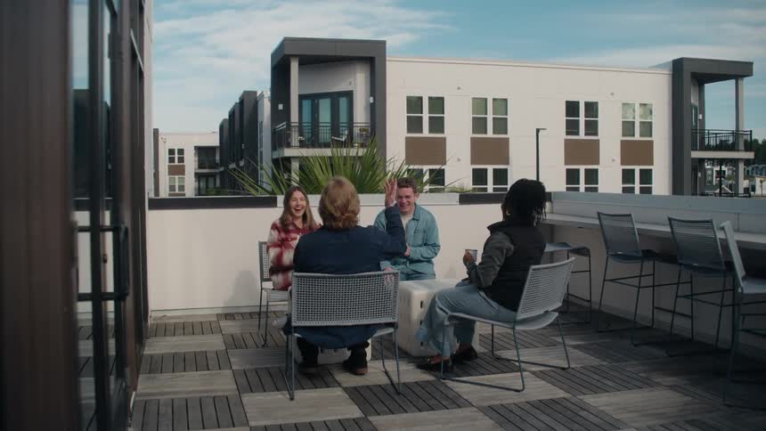 A group of four young friends hang out, talk, and share a laugh on the rooftop deck of a modern apartment building