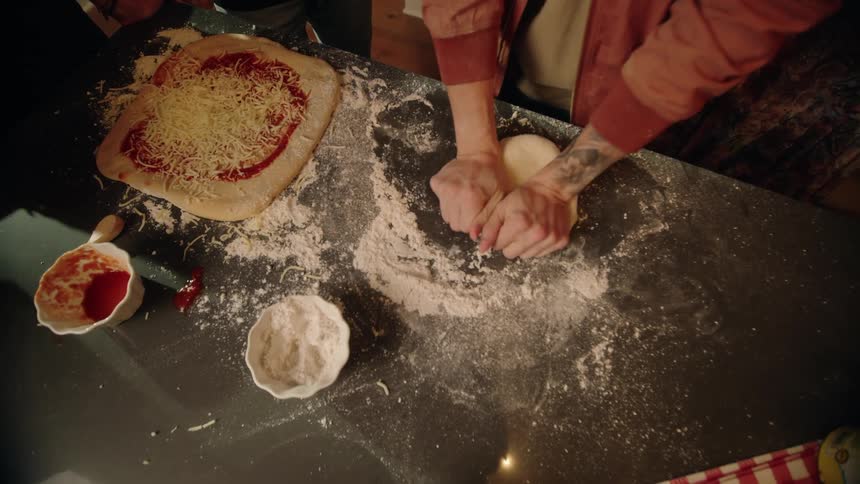 A high-angle view shows a person kneading pizza dough on a floured countertop next to a heart-shaped pizza