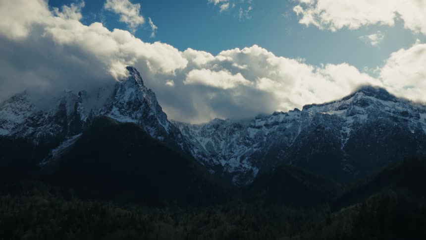 Majestic snow-capped mountains tower over a green valley under a blue sky with dramatic, rolling clouds