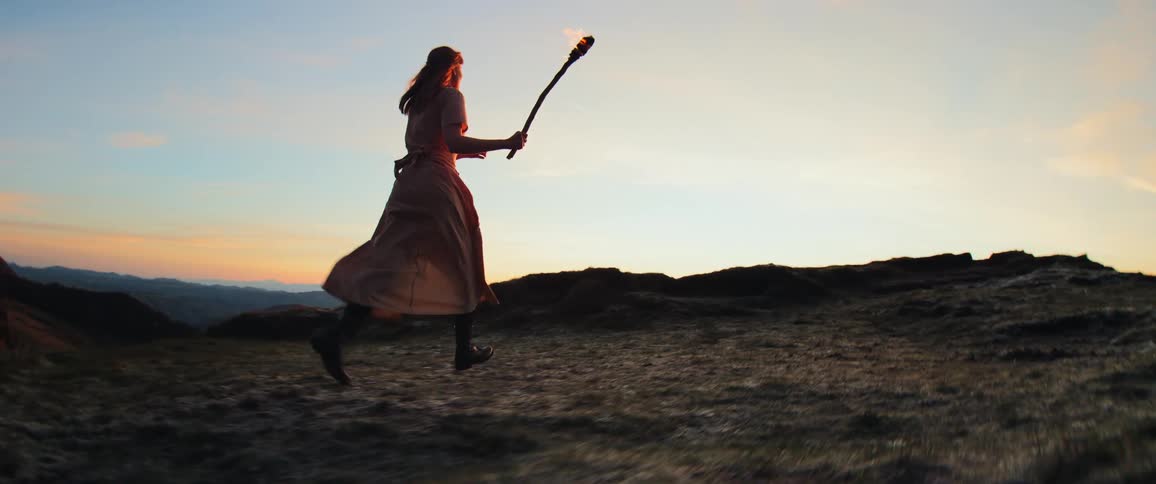 A silhouetted woman in a flowing dress runs across a mountaintop in slow motion, carrying a burning torch against the sunset sky