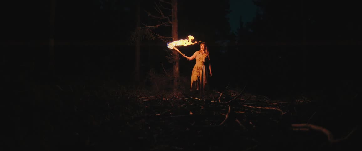 A woman in a long dress cautiously walks through a dark, eerie forest at night, holding a flaming torch to light her way