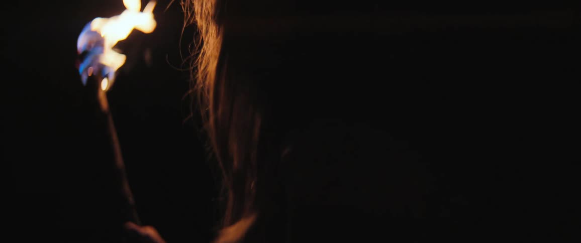 A mysterious woman holding a flaming torch walks away into the pitch-black darkness in slow motion