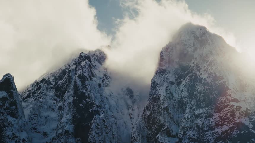 Sunlight pierces through dramatic clouds swirling around majestic, snow-covered mountain peaks in a time-lapse