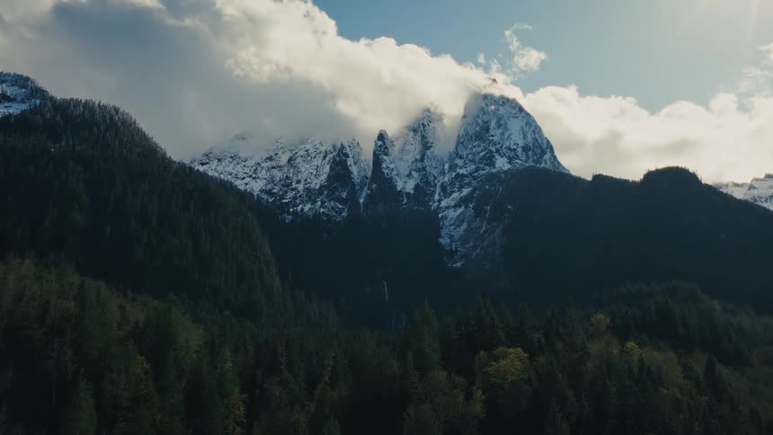 A slow aerial push-in shot of majestic snow-capped mountain peaks partially covered by clouds, rising above a vast evergreen forest