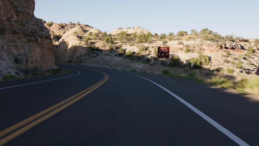 A point-of-view shot from a car driving on a winding paved road through a rocky desert canyon in the golden morning light