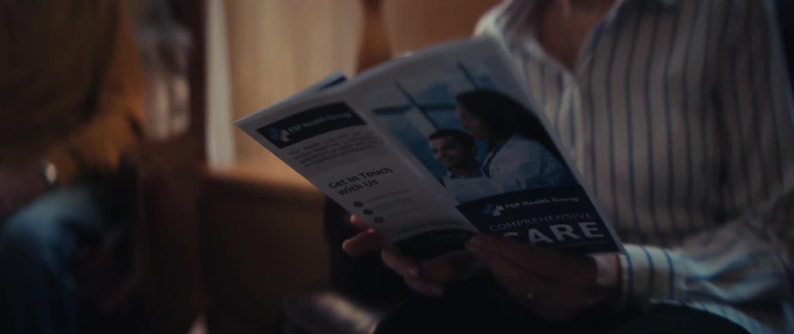 An elderly woman with a concerned expression reads a brochure for a comprehensive care health group in her living room