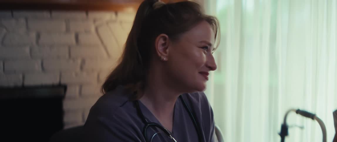 A kind female nurse in scrubs shares a warm, smiling moment with an elderly male patient in his home