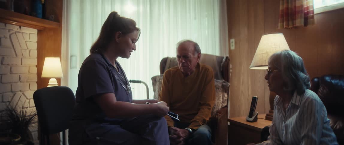 A home healthcare nurse explains a medical treatment plan to a concerned elderly couple in their living room
