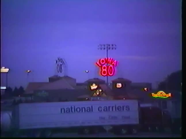 Iconic 'Iowa 80' neon sign glows brightly over a vast truck stop parking lot at dusk in this retro VHS footage