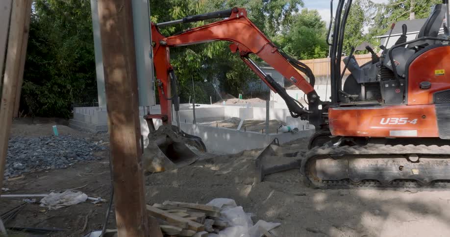 Low traverse past a mini-excavator revealing a newly poured concrete foundation on a residential construction site