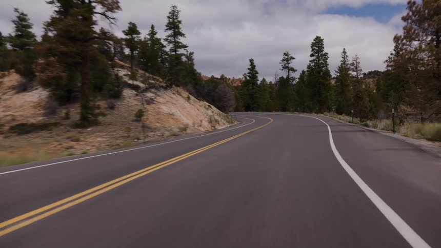 A POV shot of driving down a curvy road surrounded by evergreen trees and rocky hills under a cloudy sky