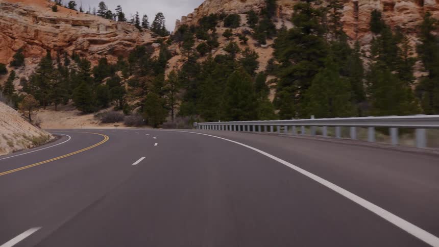 A scenic point-of-view drive on a winding mountain road surrounded by red rock canyons and lush pine trees under a cloudy sky