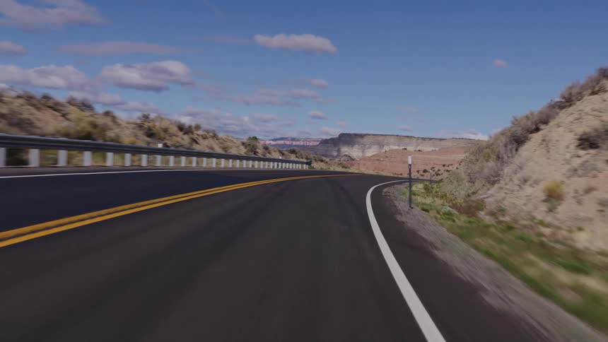 A smooth POV shot follows a winding road through the desert canyon landscape on a sunny day