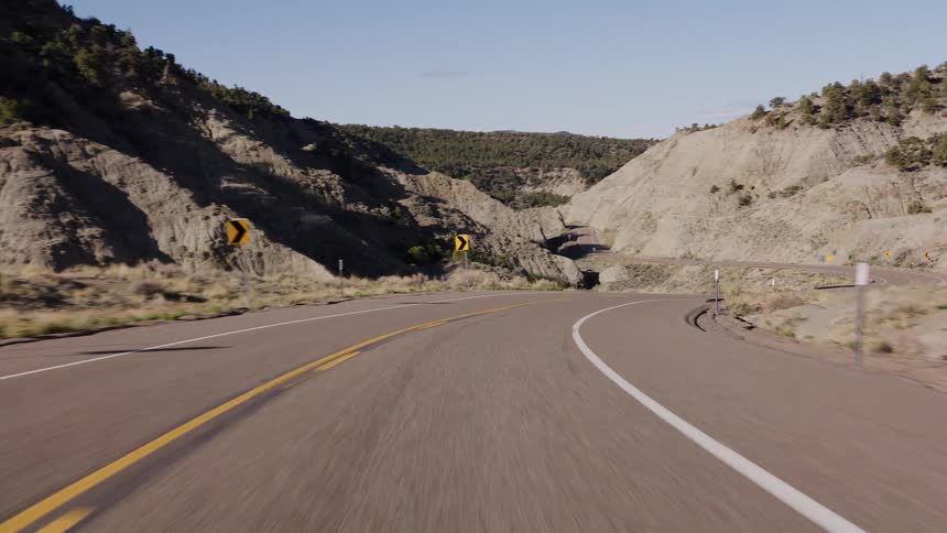 A point-of-view shot from a vehicle driving down a winding highway through a rocky, arid canyon on a sunny day