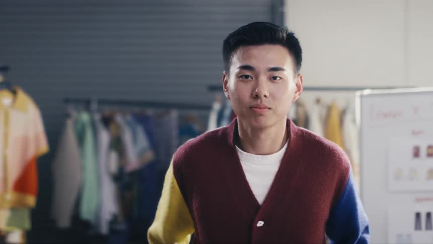 A young, stylish Asian fashion designer stands confidently in his studio, looking directly into the camera