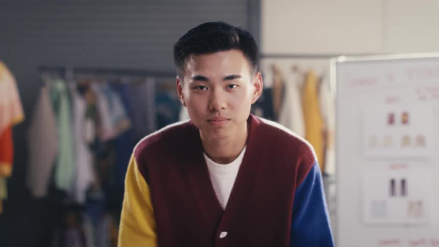 A confident young Asian man in a colorful cardigan comes into focus, looking seriously at the camera in a design studio