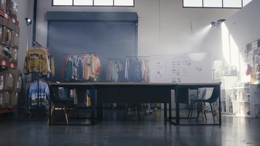 A wide, still shot of an empty fashion design studio filled with colorful clothes on racks and illuminated by a dramatic beam of light
