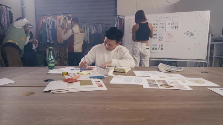Young fashion designer sketches new ideas at a large table while his diverse team works collaboratively in the background of their busy studio