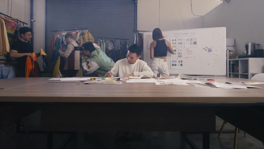 A team of young fashion designers collaborates in their studio as the camera slowly pushes in on one designer sketching new ideas